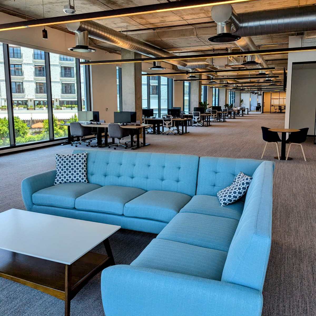 A bright office space with a blue sectional sofa and a coffee table, surrounded by empty desks and large windows.