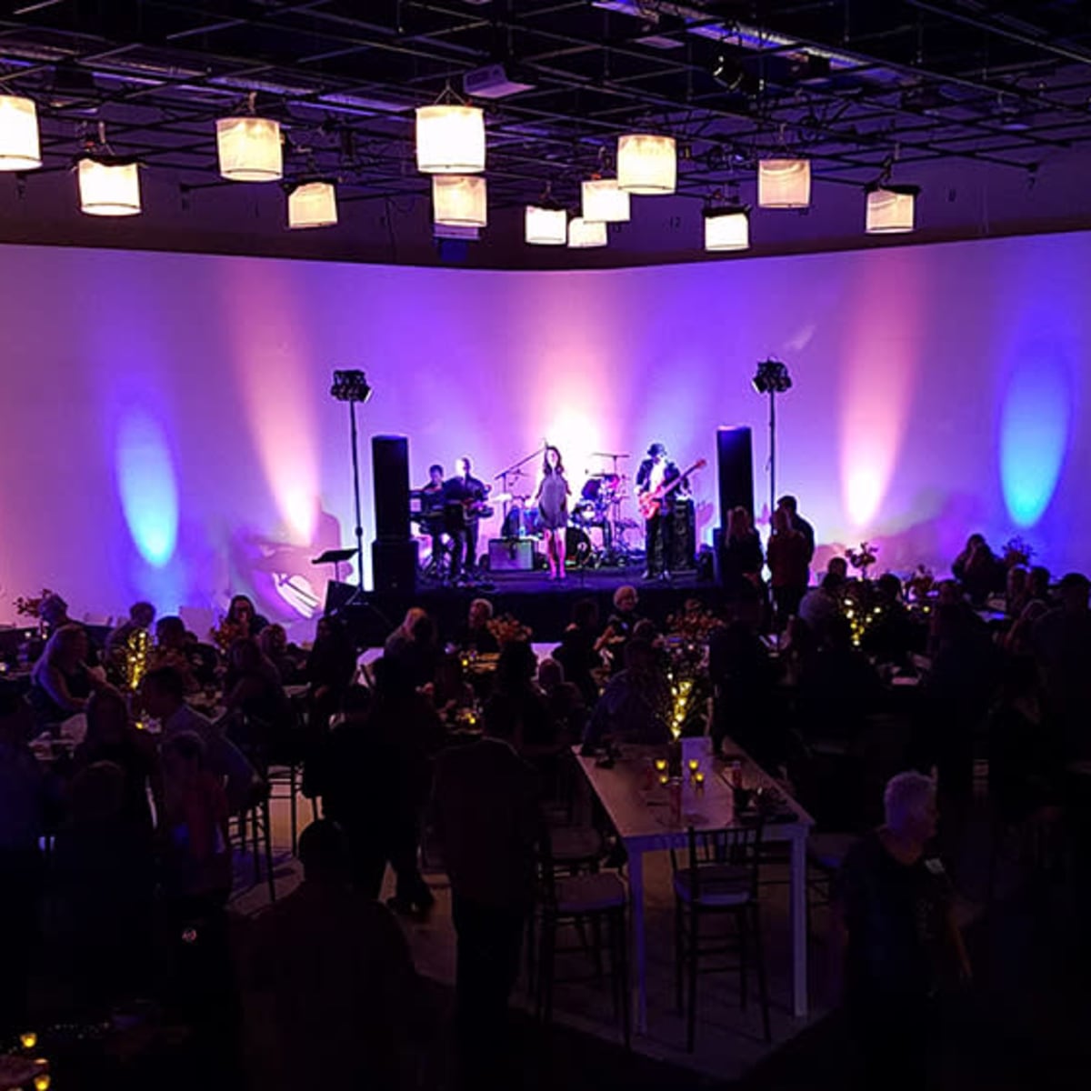 A large gathering is taking place in a venue featuring a live band on stage, illuminated by colorful lighting, with attendees mingling around tables.