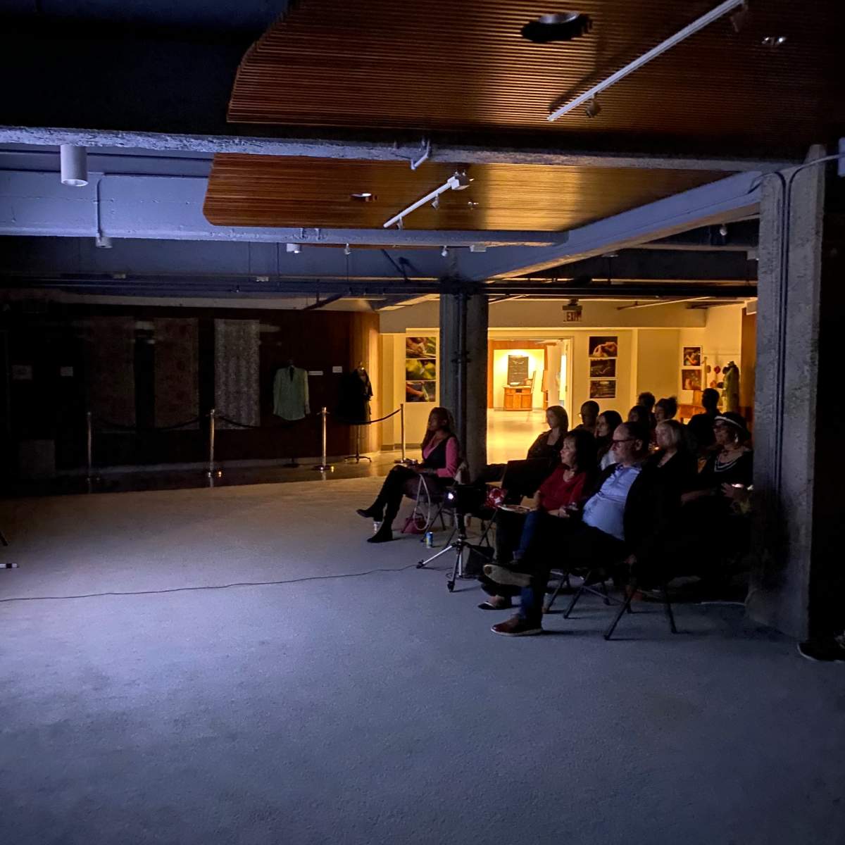 A group of people is seated in chairs facing a large screen displaying a video in a dimly lit room.