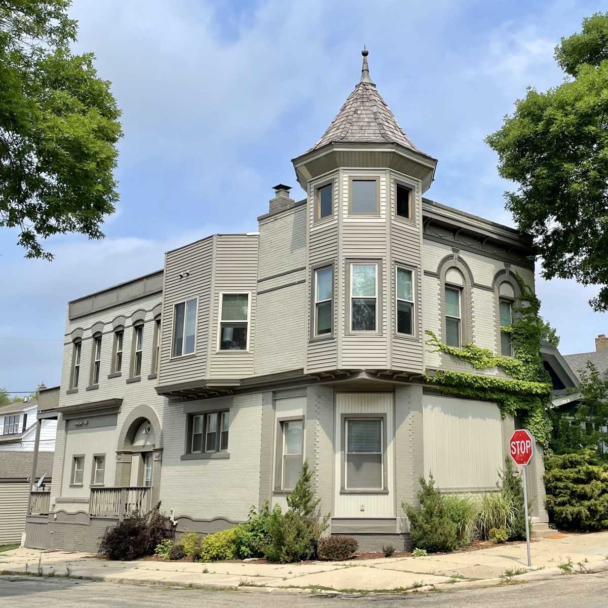 A gray, multi-story building with a distinctive turret and large windows is located at the corner of a quiet street, featuring landscaped greenery and a stop sign nearby.