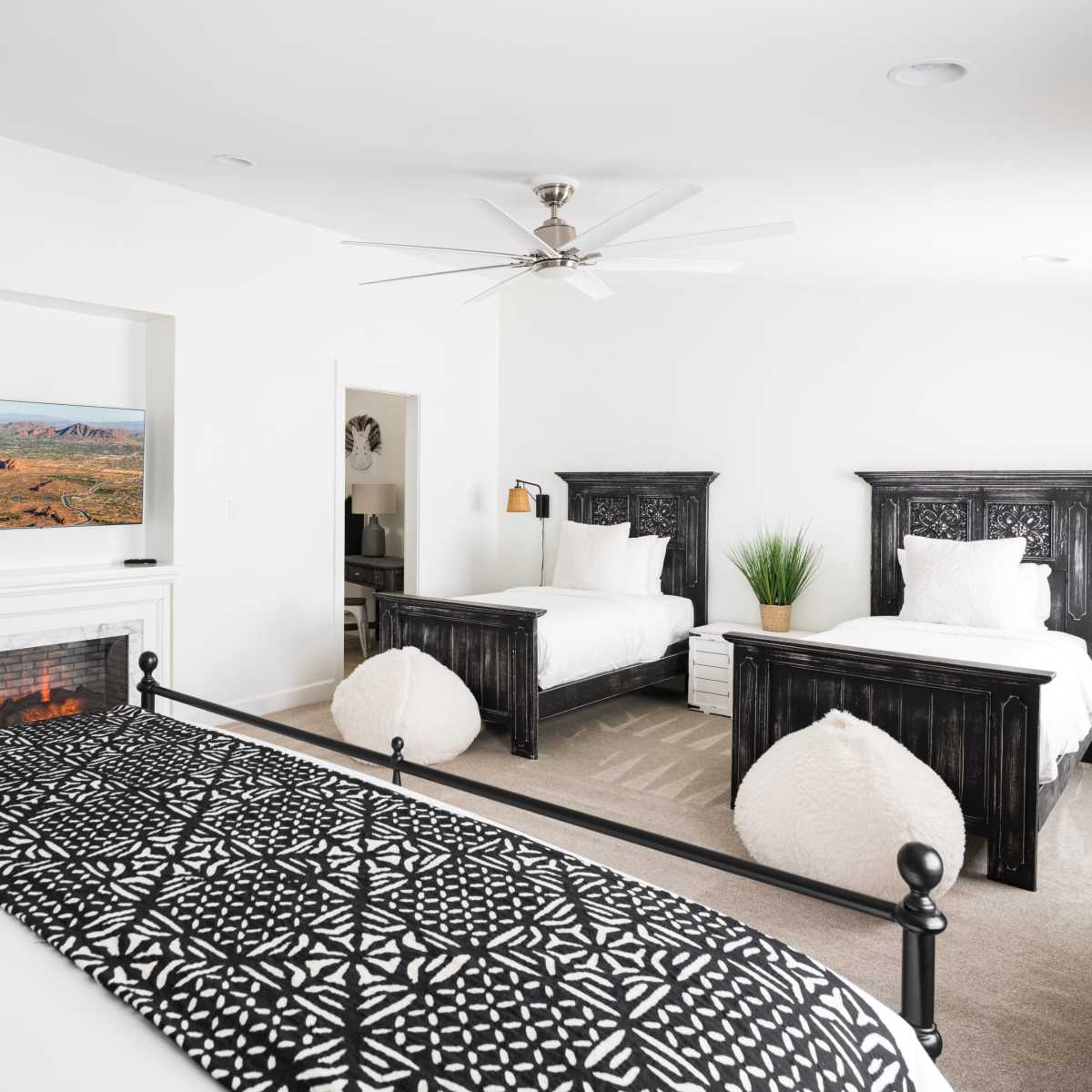 A bright bedroom featuring two twin beds with black wooden headboards, a wall-mounted TV, and a cozy seating area with a faux fireplace.