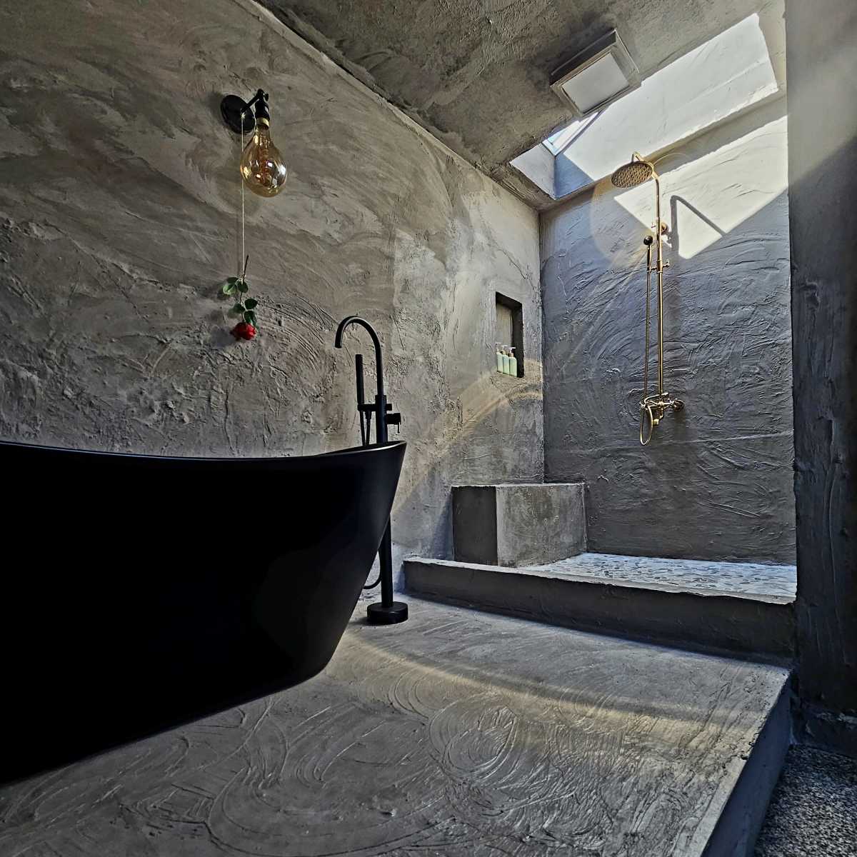 A minimalist bathroom featuring a black freestanding bathtub, a gold wall-mounted shower fixture, and a skylight above a textured concrete floor.