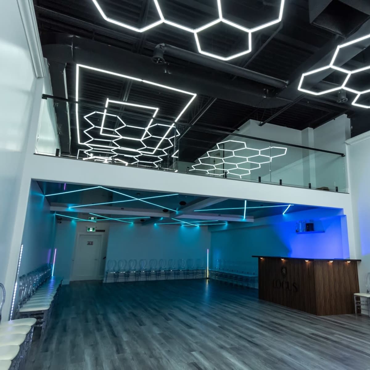 A spacious modern venue with hexagonal light fixtures on the ceiling, a sleek bar counter, and rows of transparent chairs arranged on the floor.