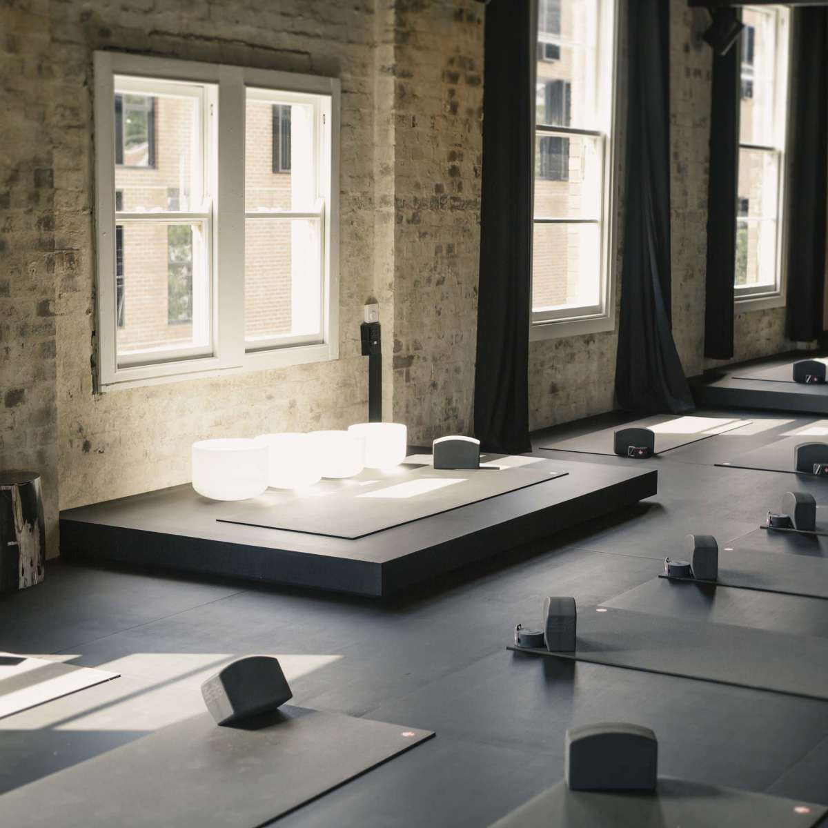 A yoga studio with several mats arranged on a black floor, illuminated by natural light coming through large windows.