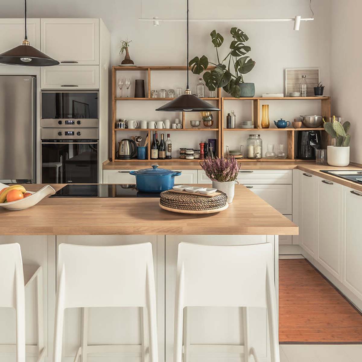 A modern kitchen with a central wooden island, three white bar stools, stainless steel appliances, and a shelf displaying various kitchen items and plants.