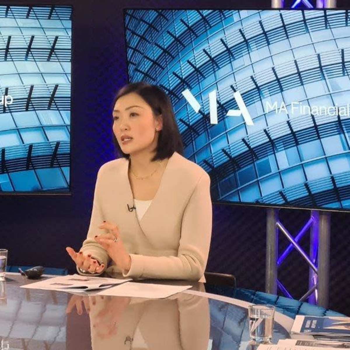 A woman is speaking at a desk with two display screens featuring a modern architectural design in the background.