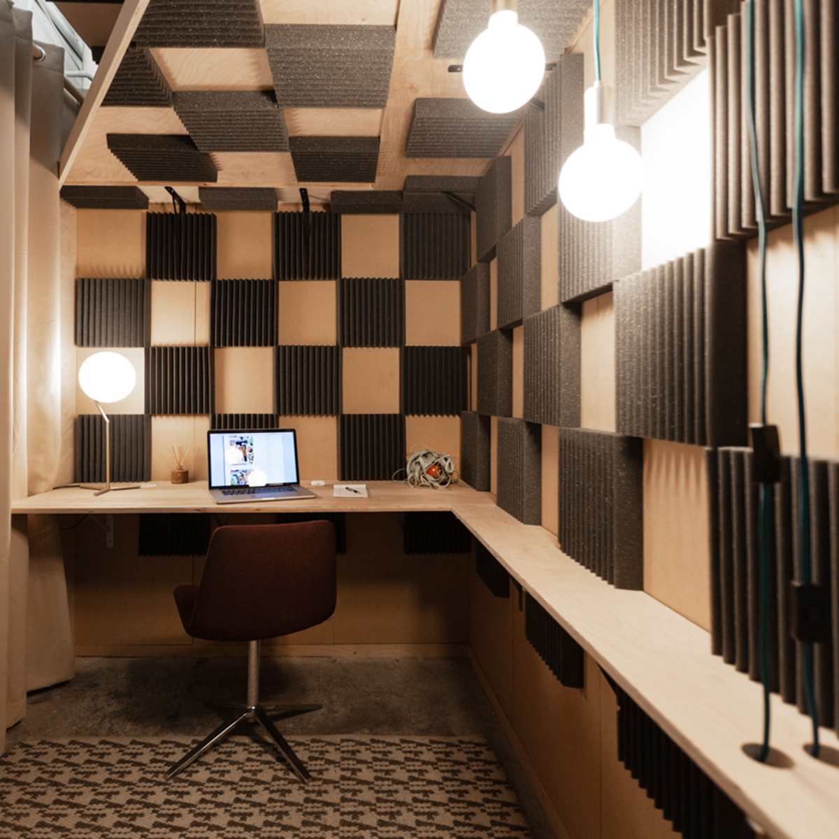 A small workspace featuring a wall of sound-absorbing panels, a desk with a laptop, and a swivel chair under soft, hanging lights.