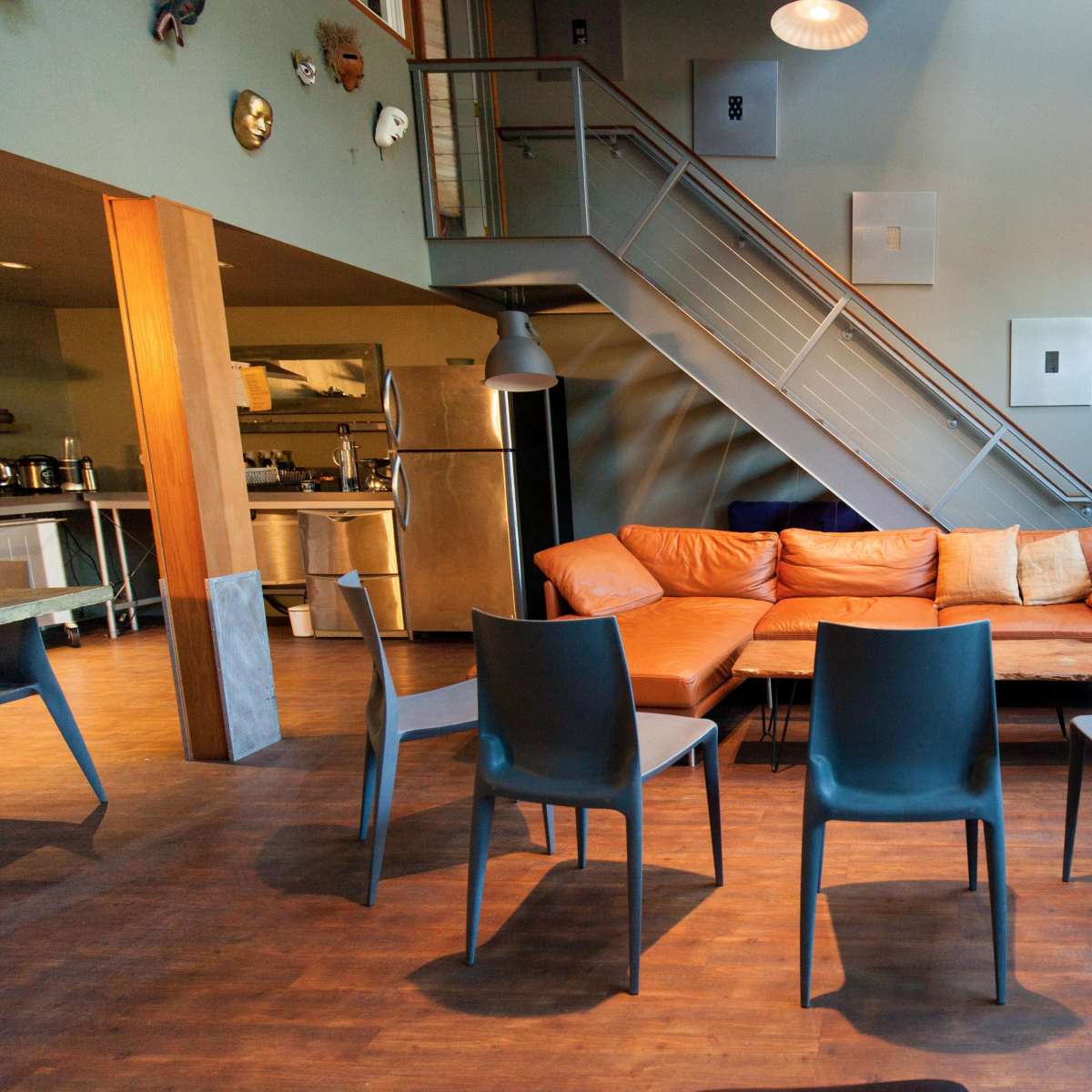 A modern living space with a staircase in the background, an orange sectional sofa, and several gray dining chairs arranged around a wooden table.