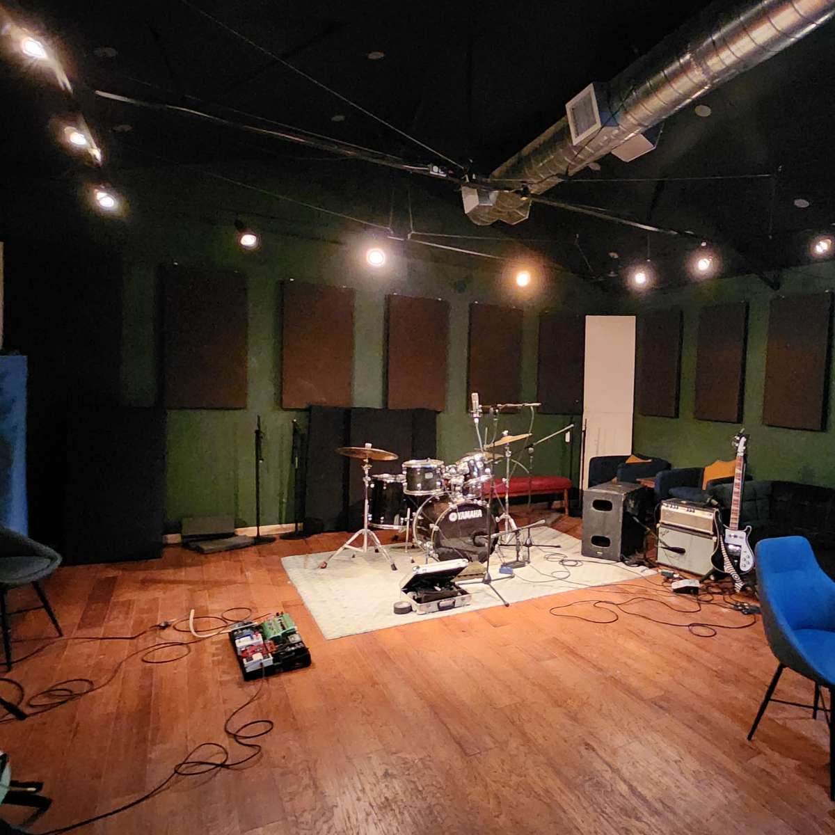A music rehearsal studio featuring a drum set, amplifiers, and sound equipment arranged on wooden flooring with soundproofing panels on the walls.