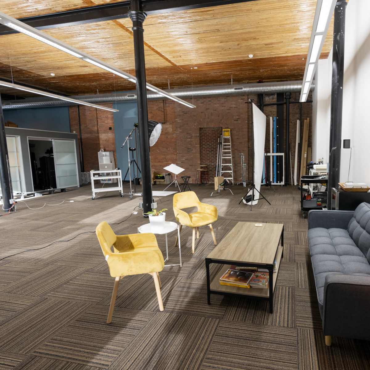 A spacious studio with a seating area featuring two yellow chairs and a gray sofa, surrounded by studio equipment and brick walls.