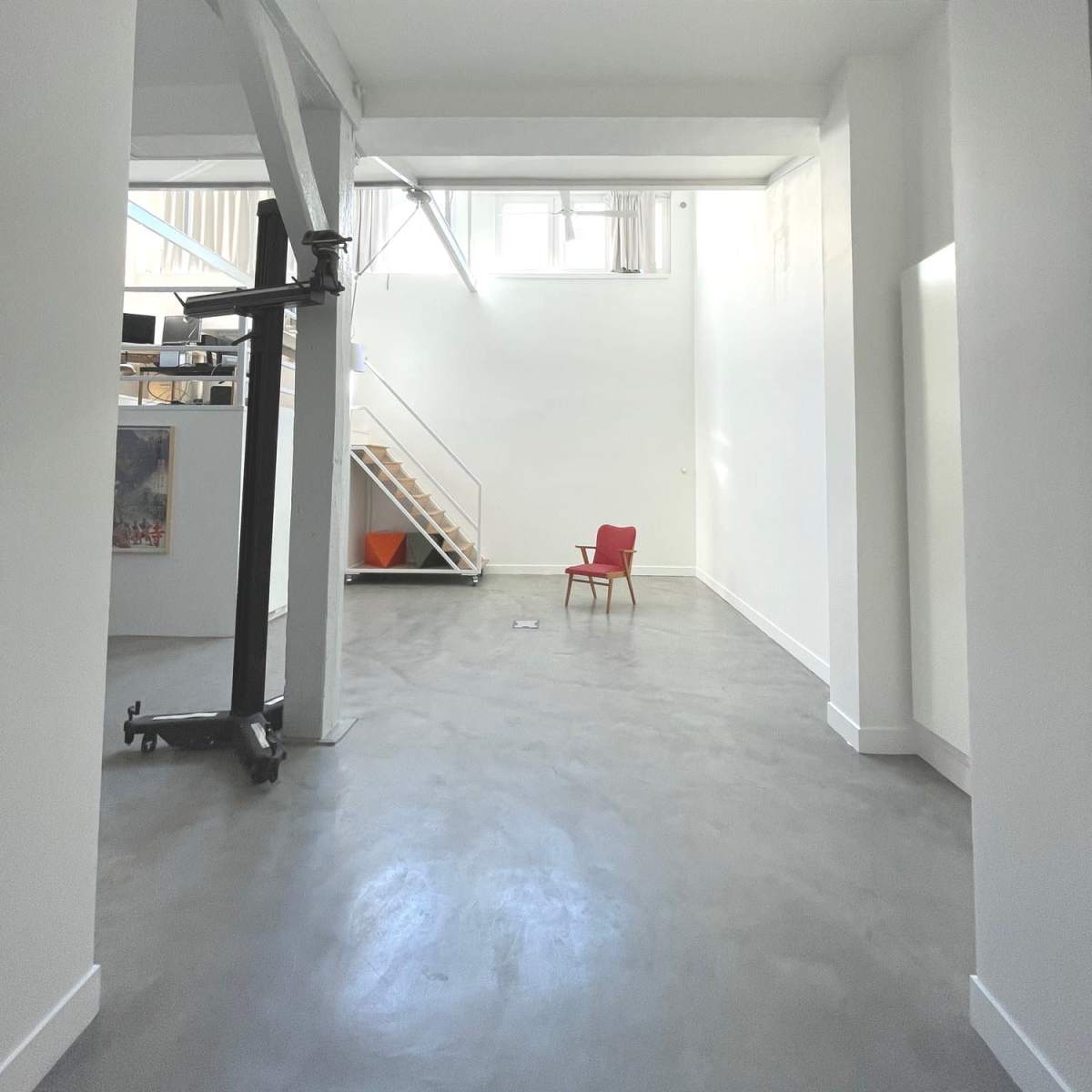 A spacious, minimalist interior with high white walls, exposed beams, and a single red chair positioned in the center.