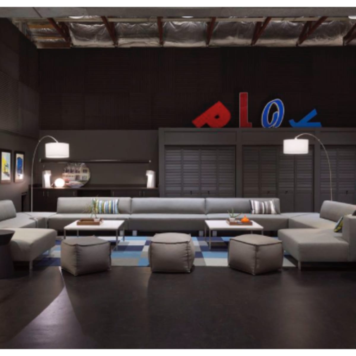 A modern lounge area with a gray sectional sofa, several small ottomans, and floor lamps, all arranged around a patterned rug in a room featuring dark walls and colorful decorative letters.