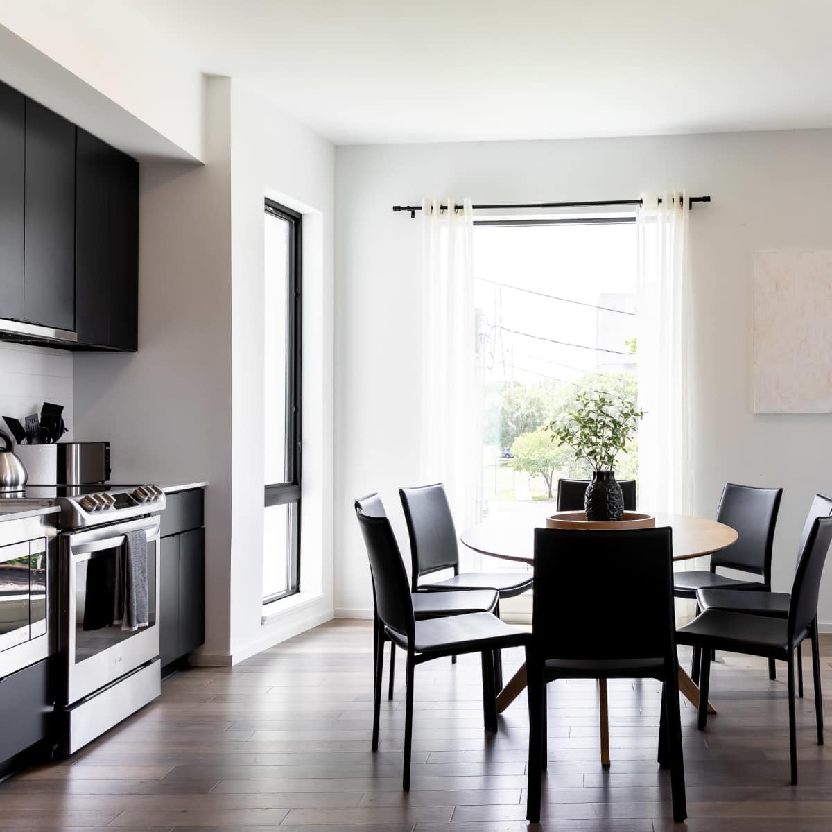 A dining area features a round table surrounded by four black chairs, with large windows letting in natural light.