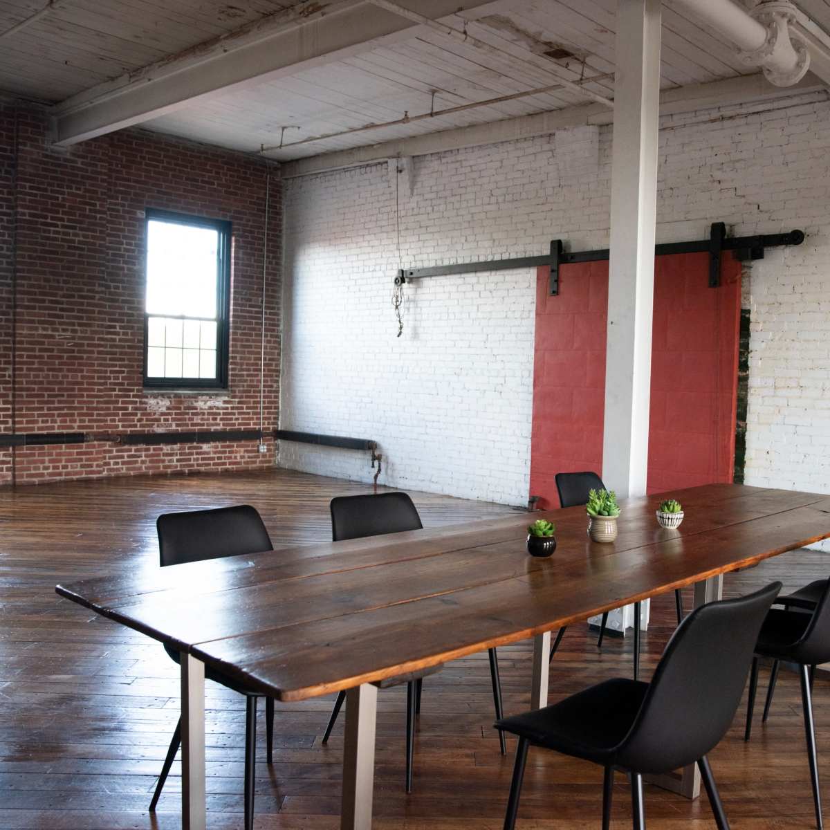 A large wooden table with chairs surrounds it in an industrial-style room featuring brick walls and red sliding doors.