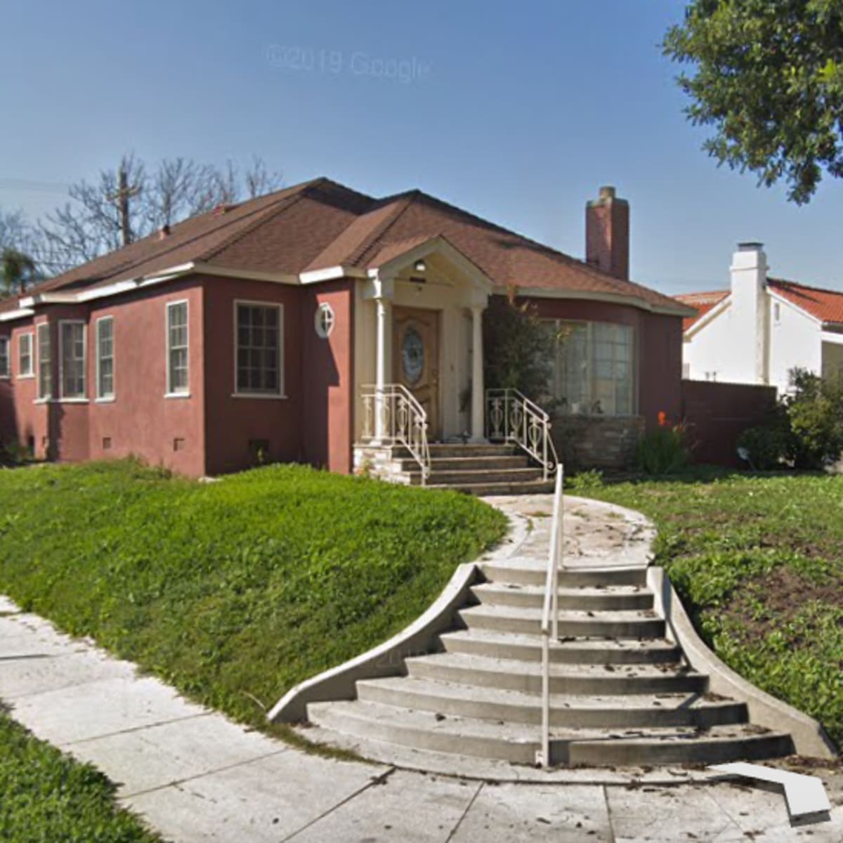 A single-story house with a red exterior and a prominent staircase leading to the front entrance, situated next to a white house with a similar structure on a grassy corner lot.