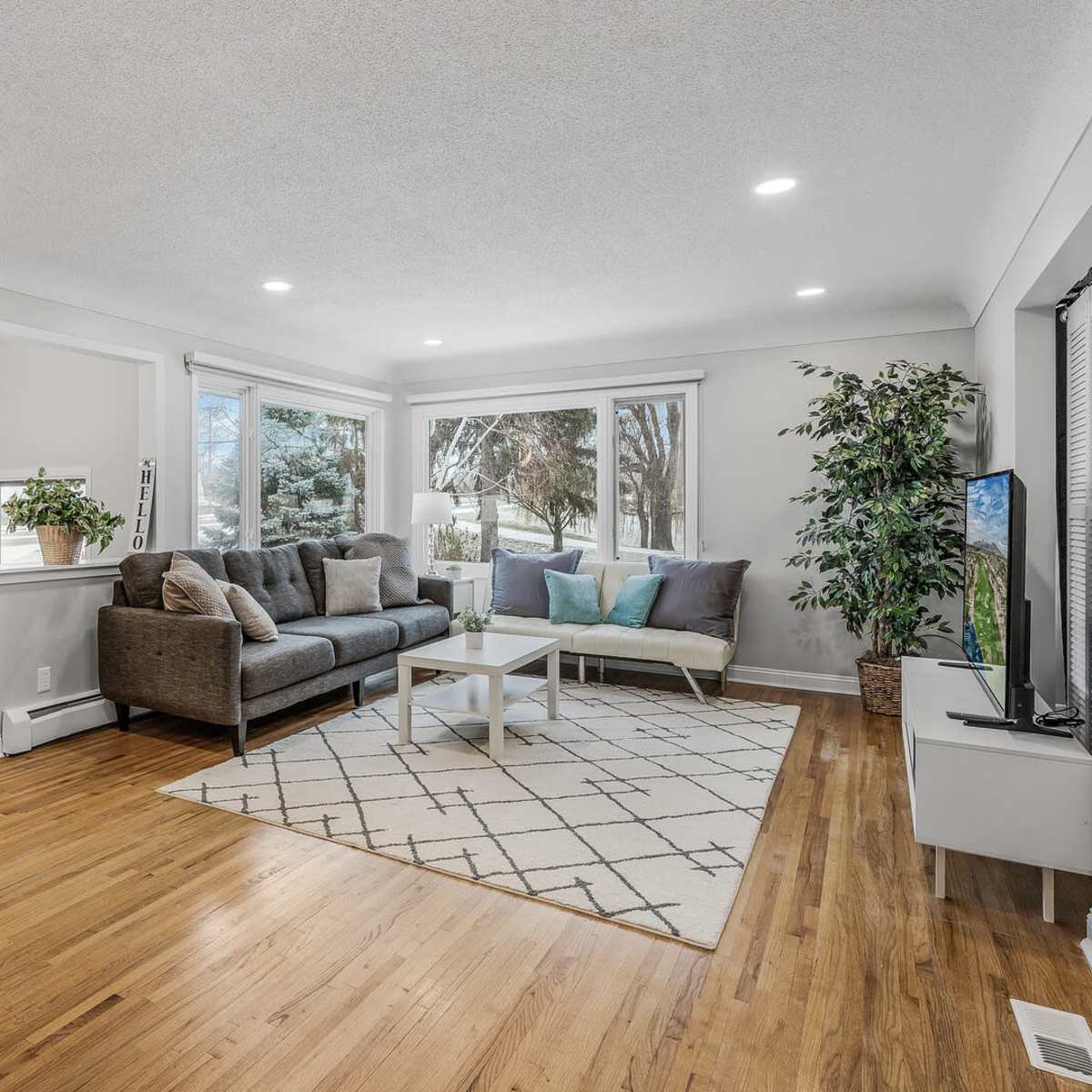 A modern living room features a gray sectional sofa, a light rug with a geometric pattern, and large windows that provide natural light.