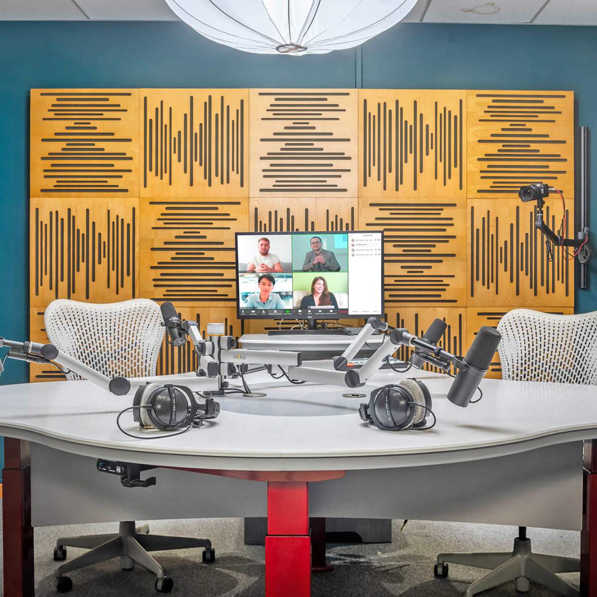 A modern recording studio with two chairs, multiple microphones, headphones, and a display screen showing remote participants.