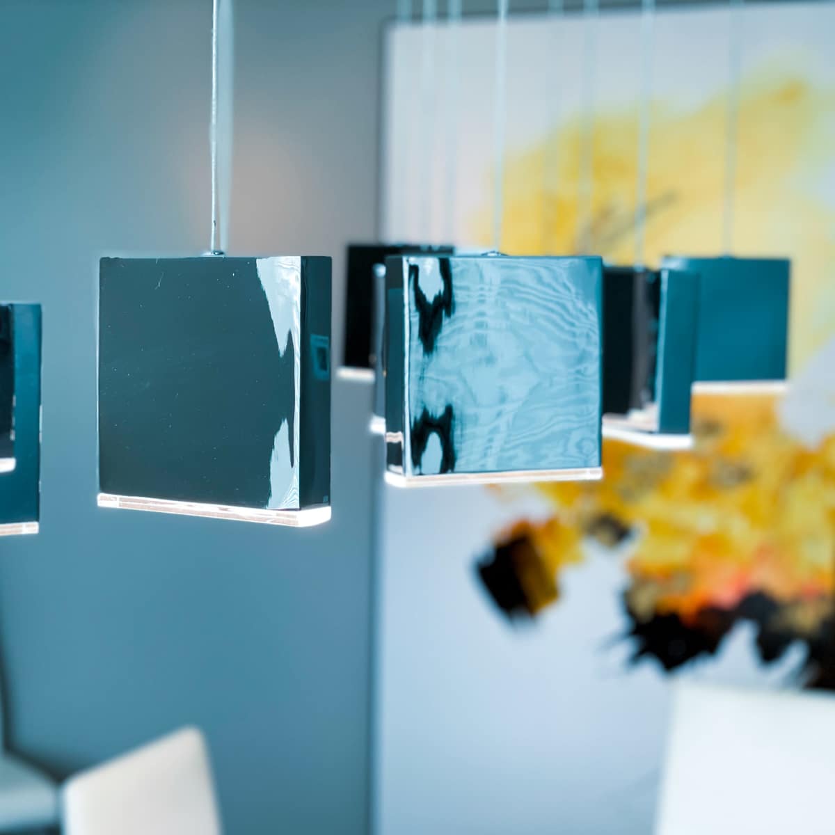 Several modern, square hanging light fixtures above a table, with an abstract yellow and black painting in the background.