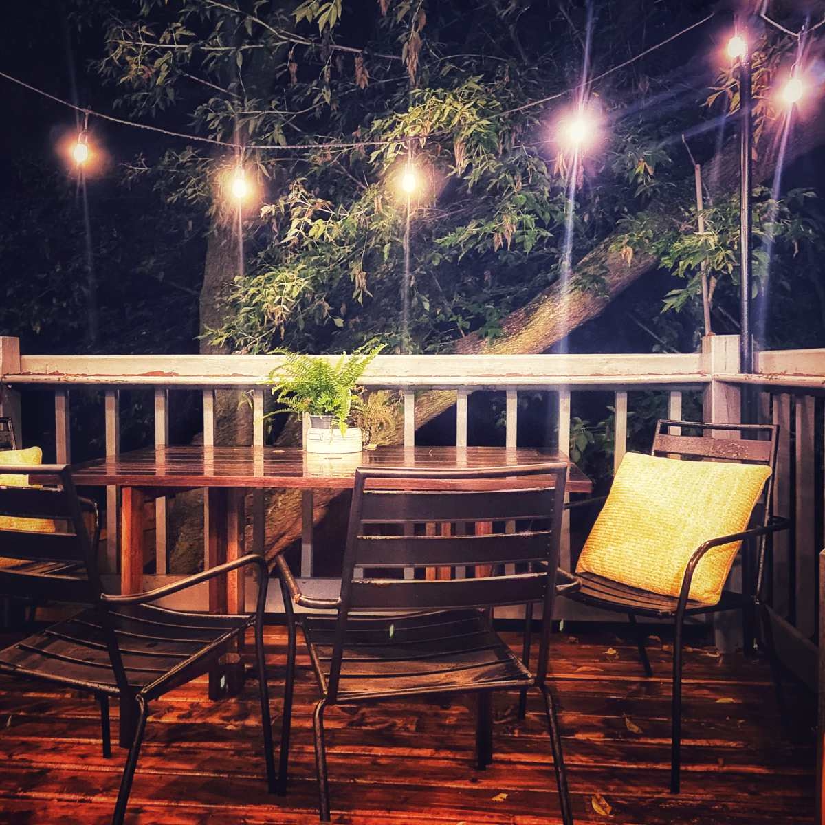 A cozy outdoor deck features a round table surrounded by four black metal chairs, illuminated by string lights overhead.