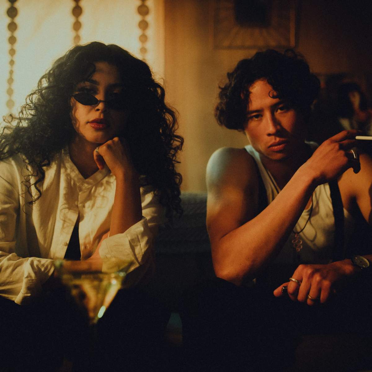 A man and a woman sit together on a couch in a dimly lit room, with the woman wearing sunglasses and the man holding a cigarette.