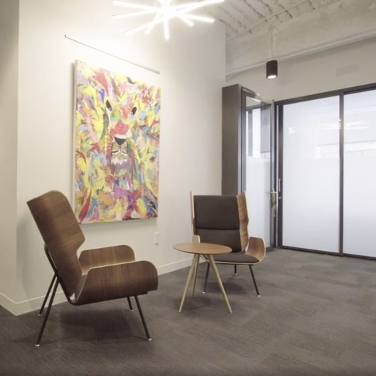 A modern office interior featuring two chairs and a small table in a brightly lit area, with an artwork on the wall and a hallway visible in the background.