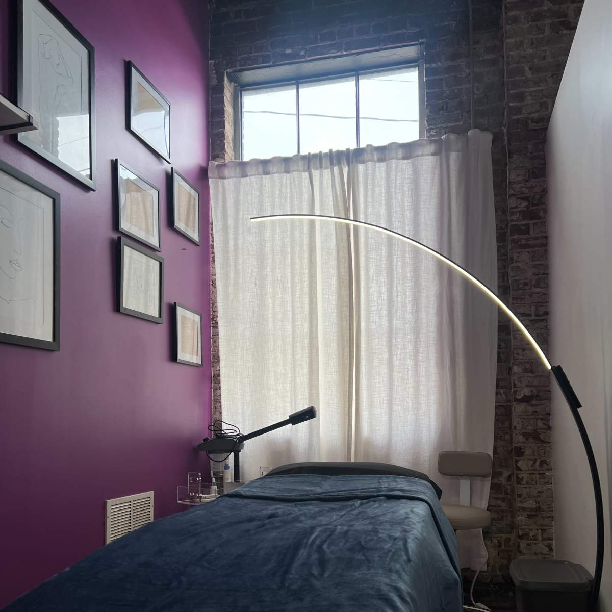 A room with a purple wall featuring framed artwork, a massage table covered with a blue blanket, and a tall lamp beside a chair near a window with sheer curtains.