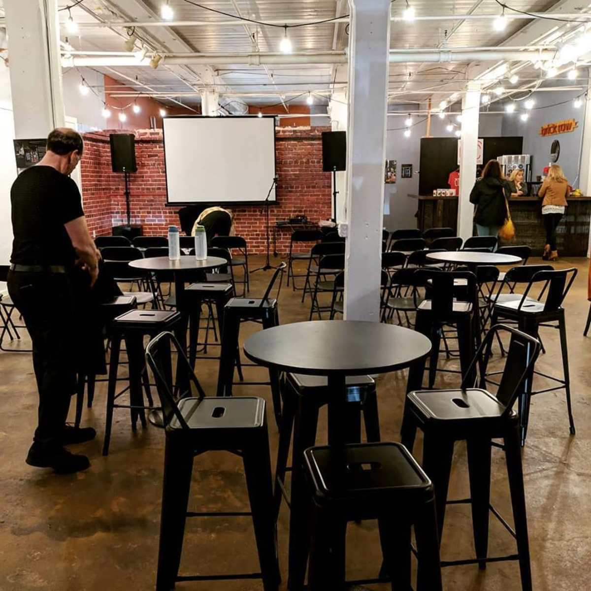 A spacious event venue is set up with black tables and chairs arranged for an upcoming gathering, with a screen and bar area in the background.