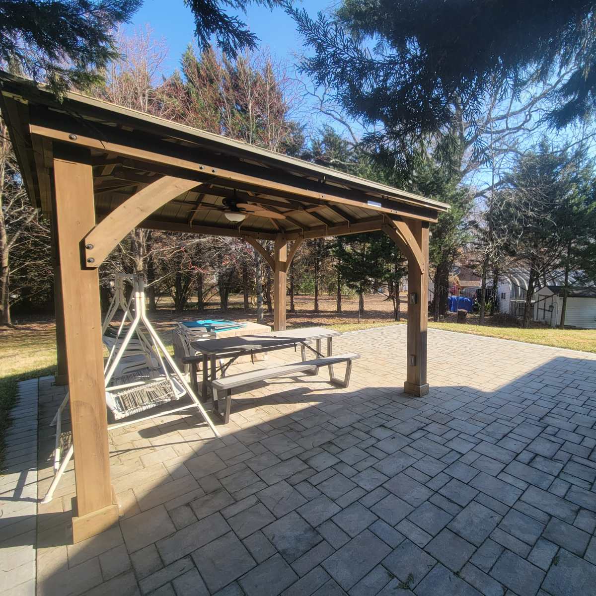 A patio area featuring a wooden gazebo with a table and chairs, surrounded by trees and a grassy lawn.