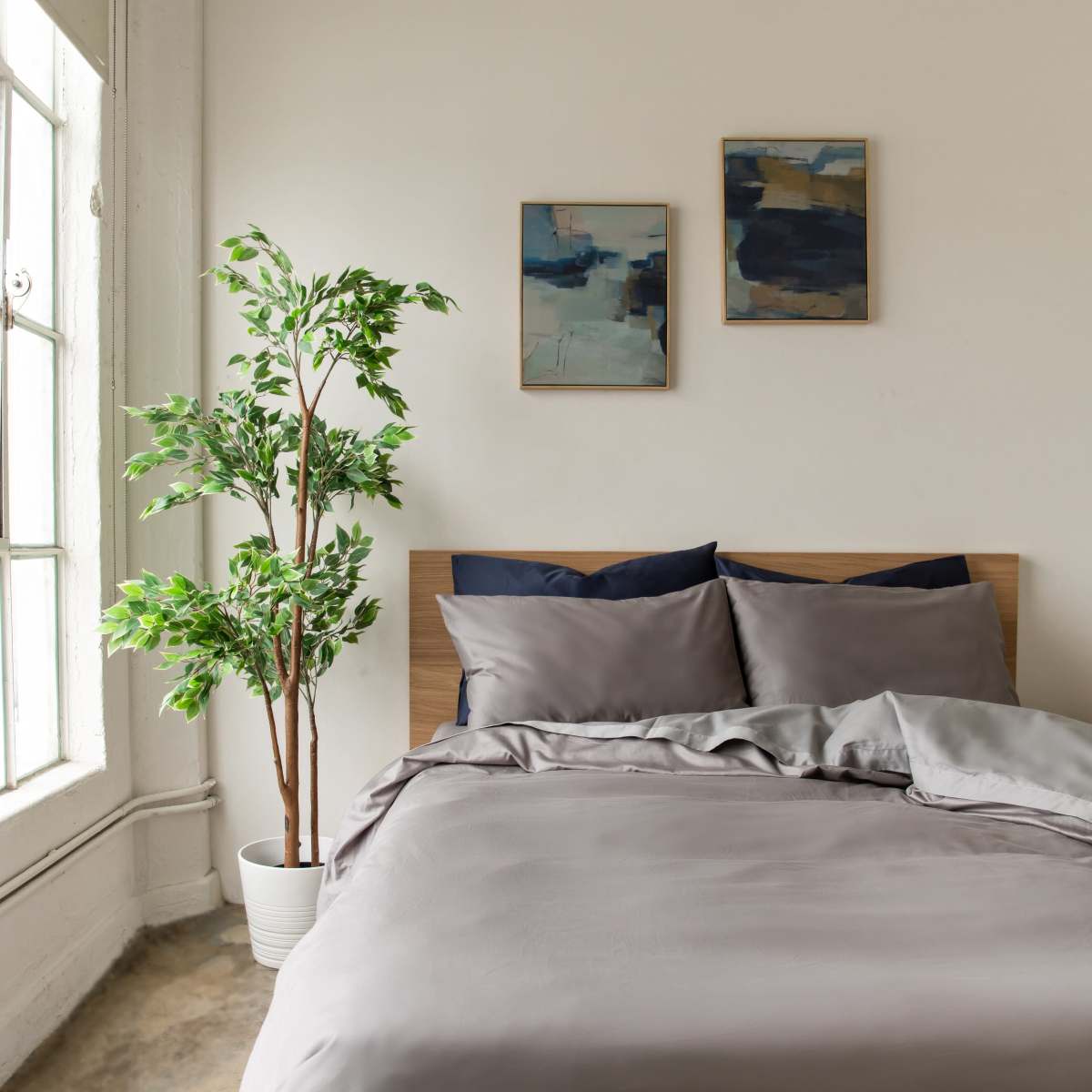 A neatly made bed with gray linens is positioned against a wall, accompanied by two framed artworks and a potted plant near a large window.