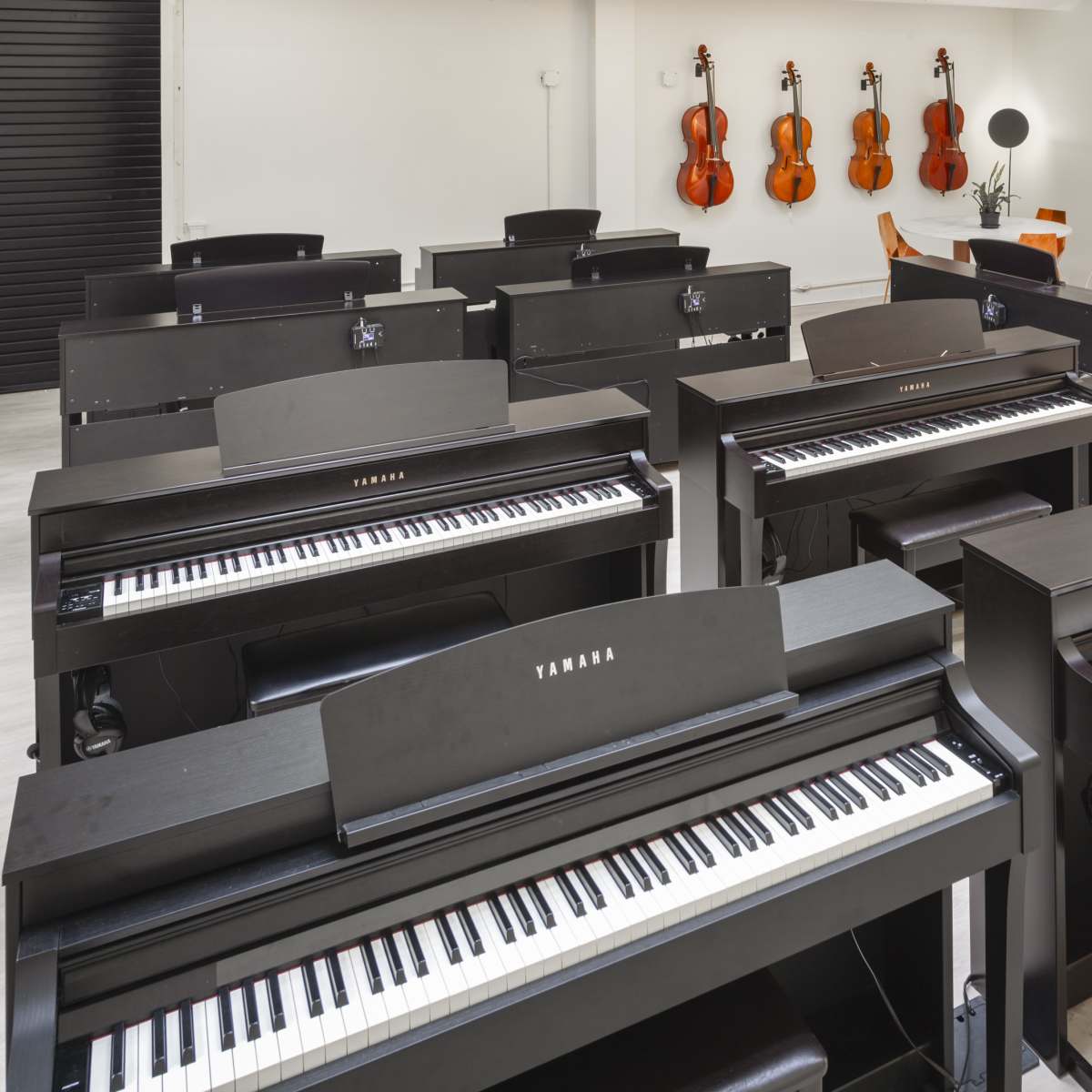 A music store interior featuring multiple black digital pianos arranged in rows, with violins hanging on the wall in the background.
