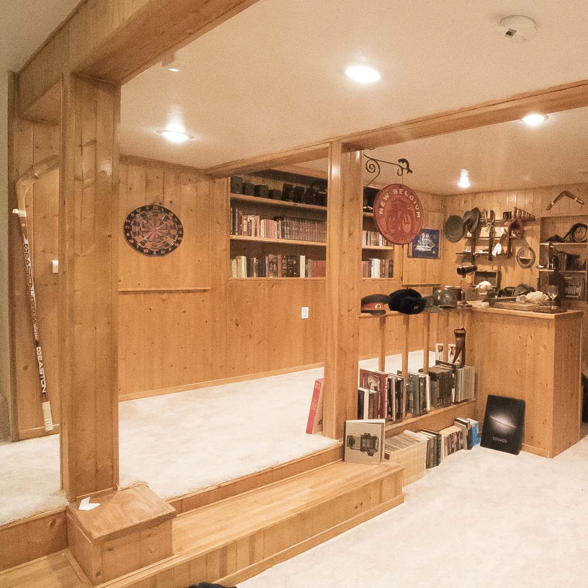 A wooden-paneled basement with a small kitchen area, shelves filled with books and decorative items, and various musical instruments hanging on the wall.