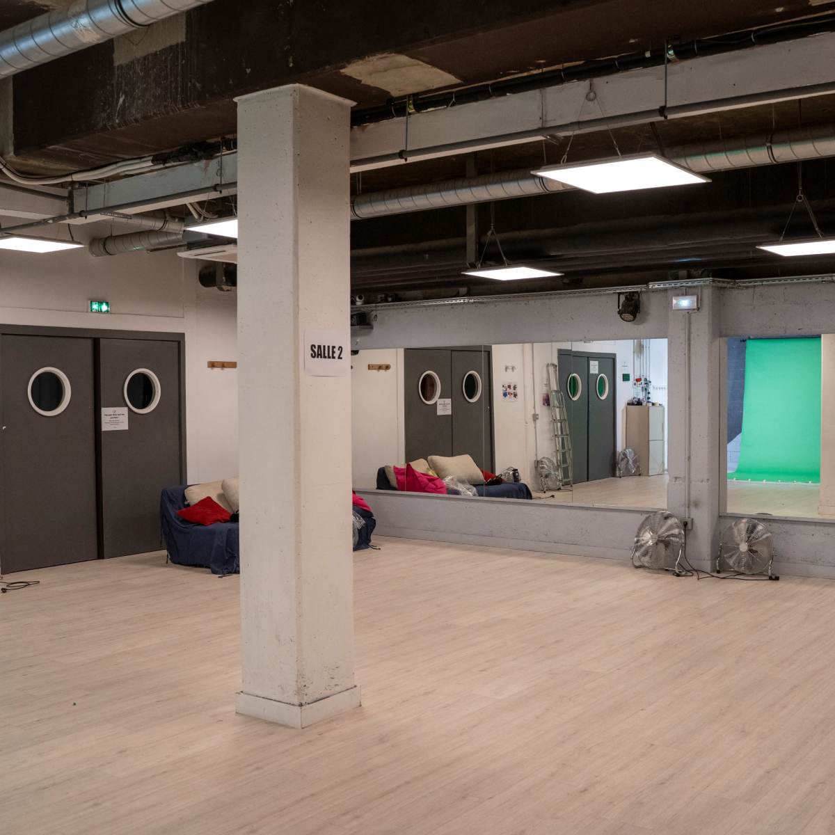 A spacious room with mirrors, a staircase, and a green screen area, featuring seating along one wall.