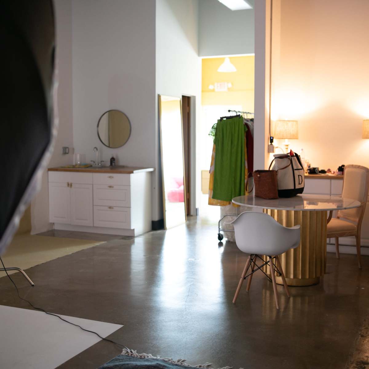 A studio space featuring a bathroom area, a clothing rack, a desk with a chair, and soft lighting.