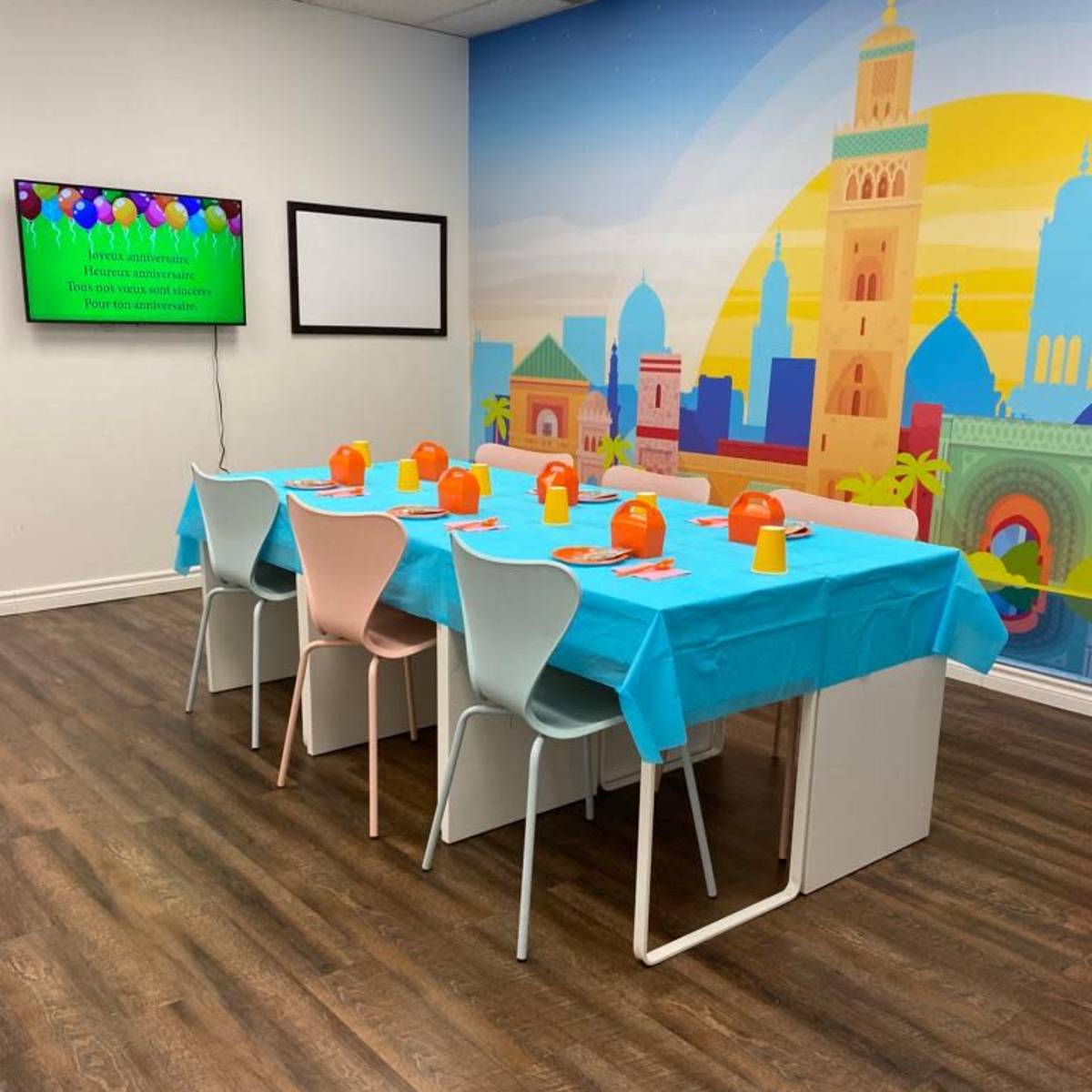 A brightly decorated party room features a table covered with a turquoise tablecloth, set with colorful plates and cups, alongside a large mural depicting a scenic landscape.