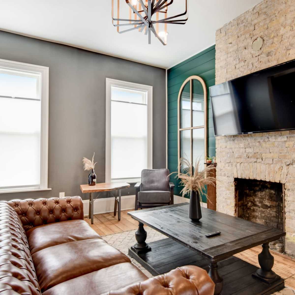 The room features a leather sofa, a stone fireplace, a mounted television, and green accent walls with large windows.