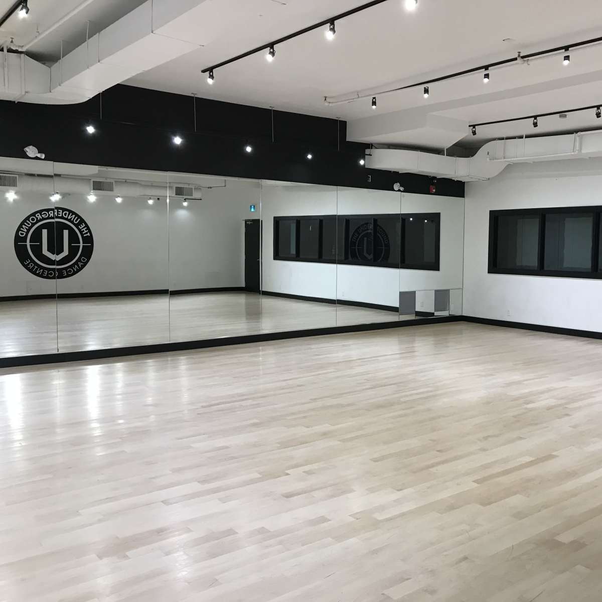 An empty dance studio with wooden flooring, mirrors on one wall, and large windows on another wall.