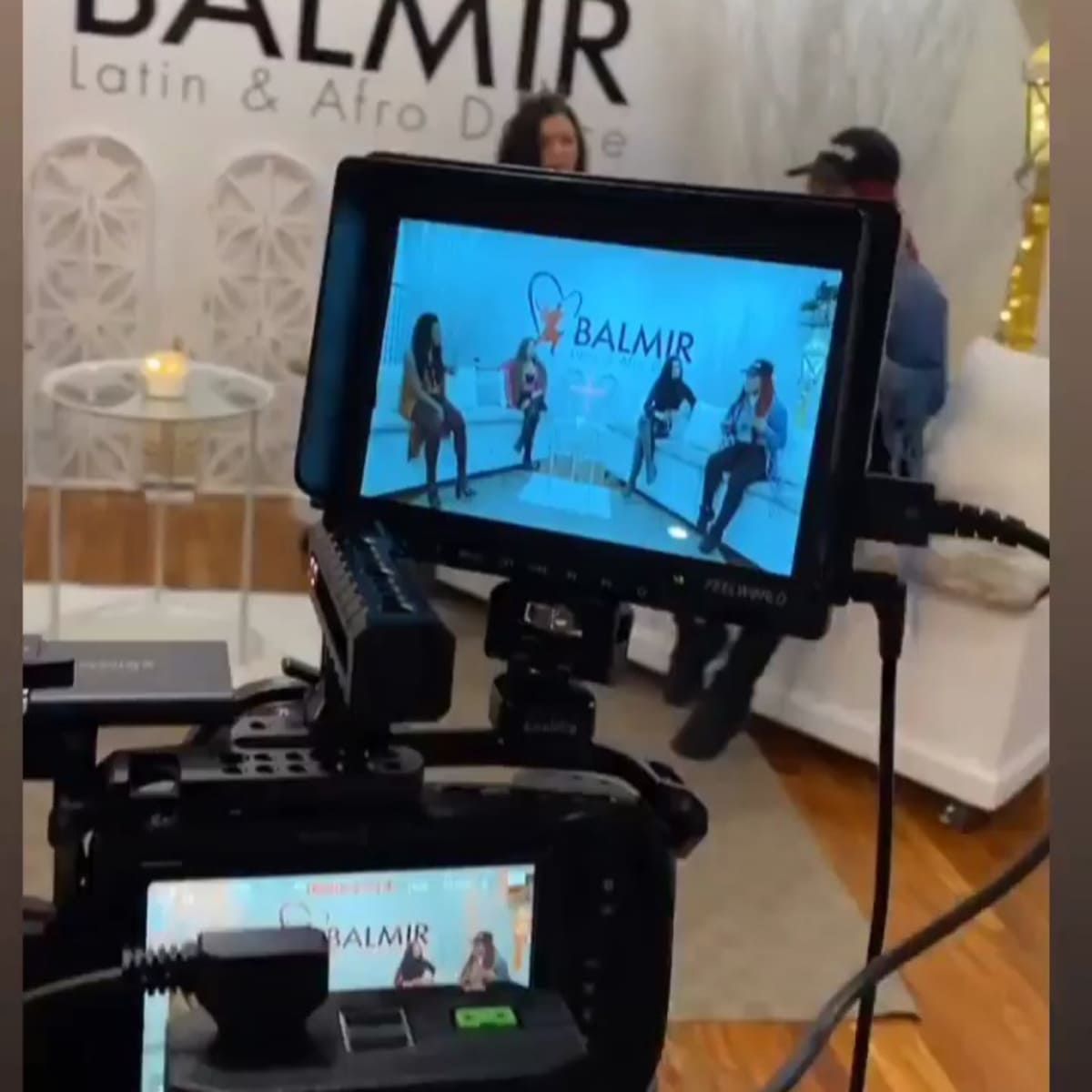 A video production setup featuring a camera focused on a group discussion taking place in a stylish studio with a "BALMIR Latin & Afro Dine" backdrop.