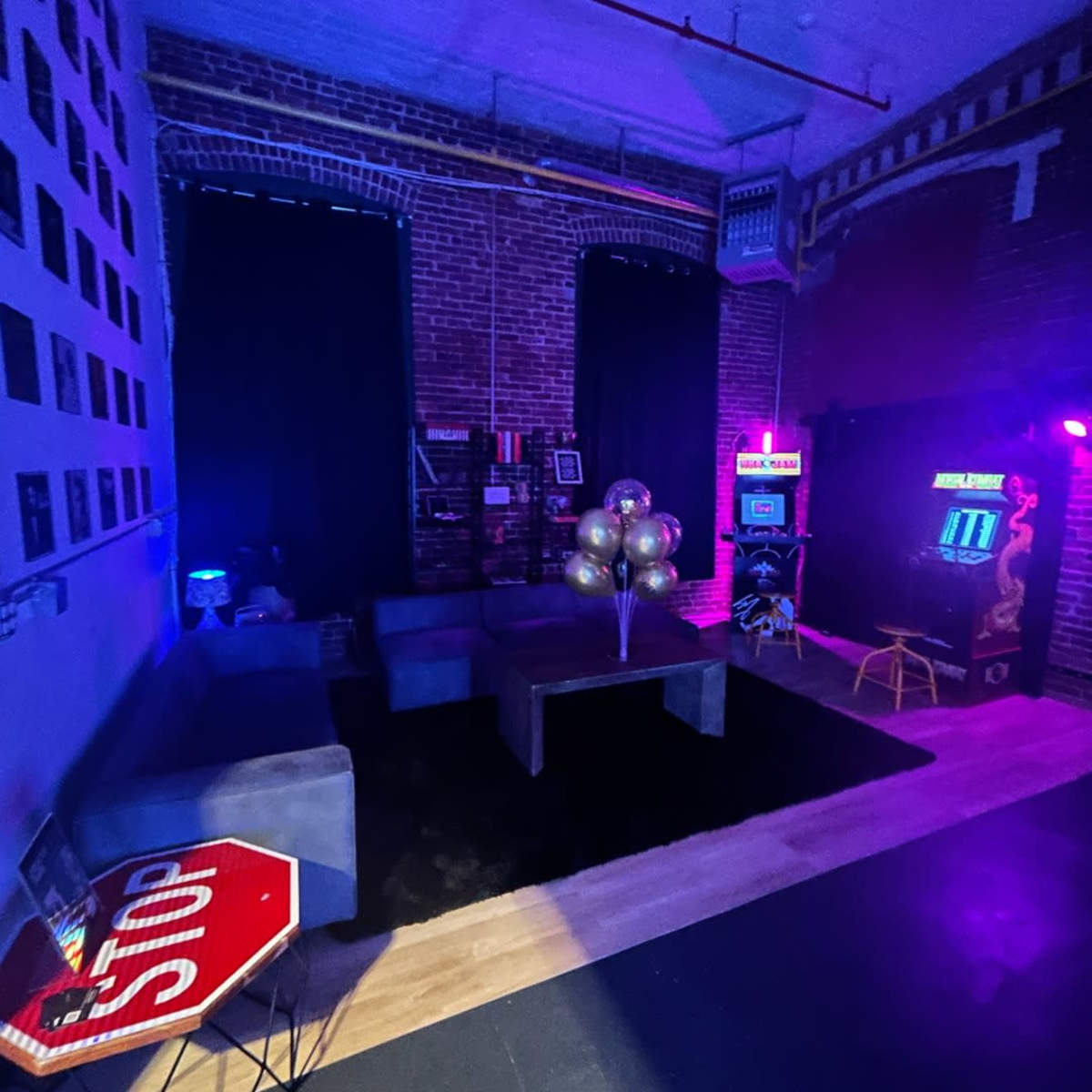 A dimly lit lounge area with a black carpet, a coffee table, gray sofas, a stop sign, and an arcade machine in the background.