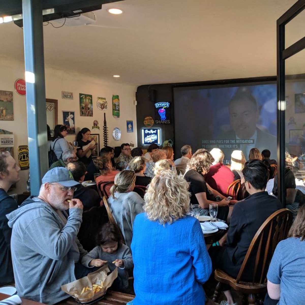 A crowd of people sits in a cafe, watching a large screen displaying a presentation.