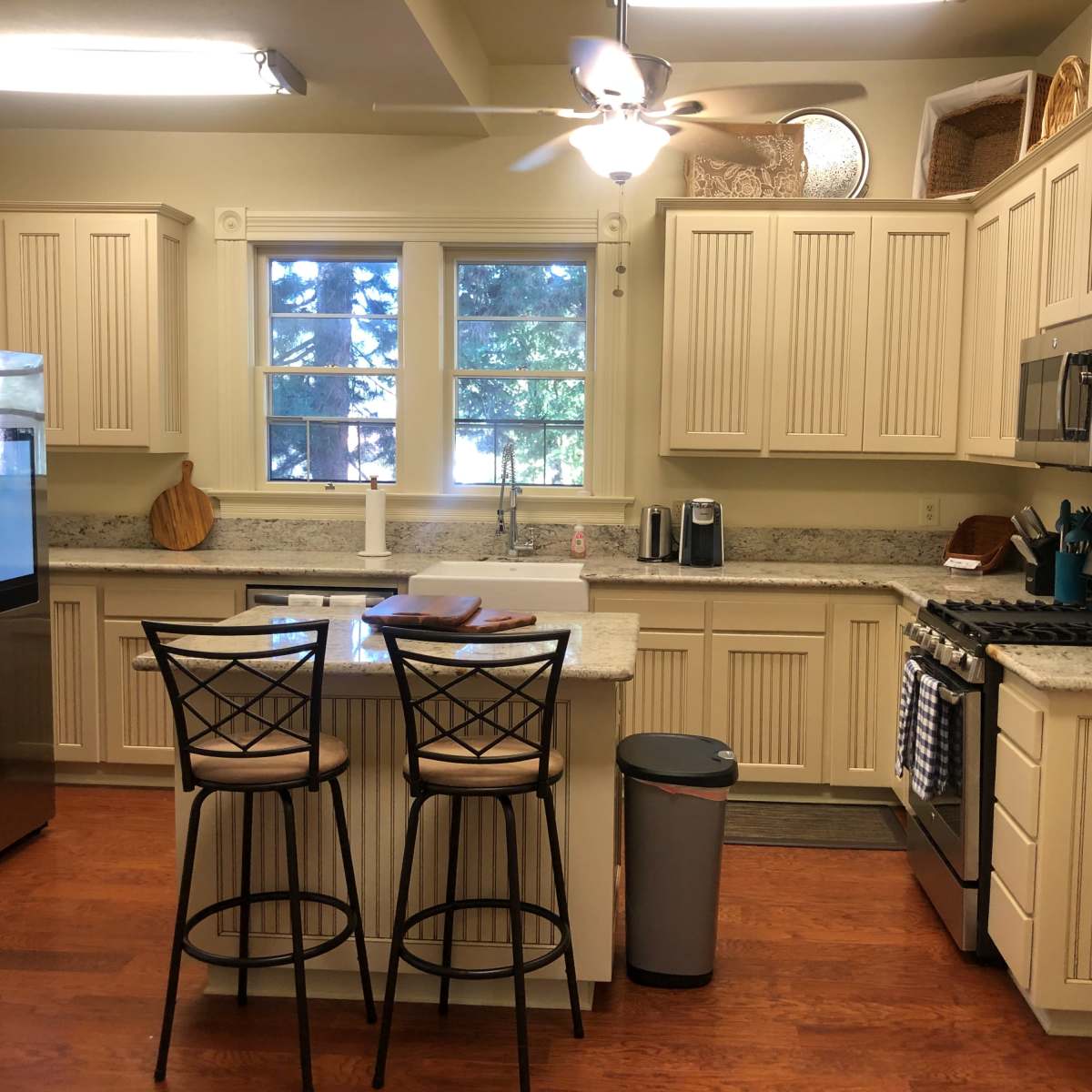 A modern kitchen features light-colored cabinetry, a granite countertop island with two bar stools, and stainless steel appliances.