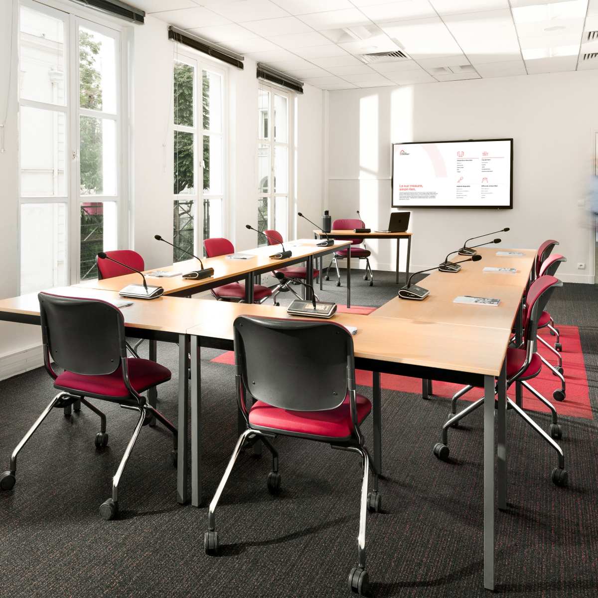 A modern conference room features a large table surrounded by red chairs, with a display screen on the wall and large windows allowing natural light.
