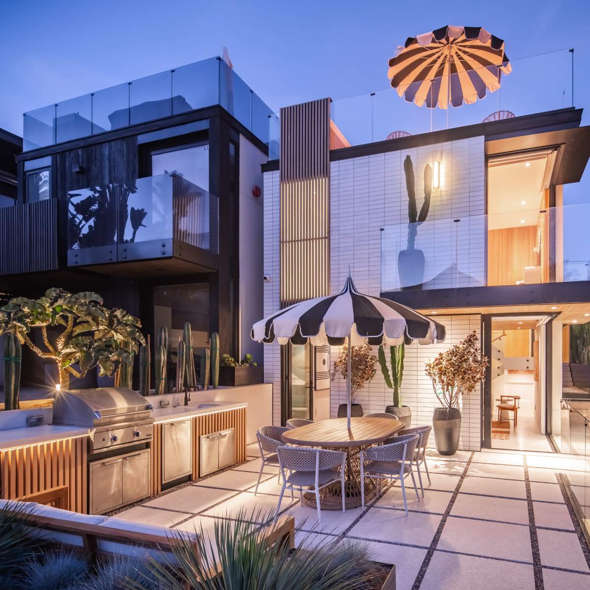 A modern two-story house features outdoor dining furniture, a grill area, and tall cactus plants in a well-lit backyard with a distinctive striped umbrella.