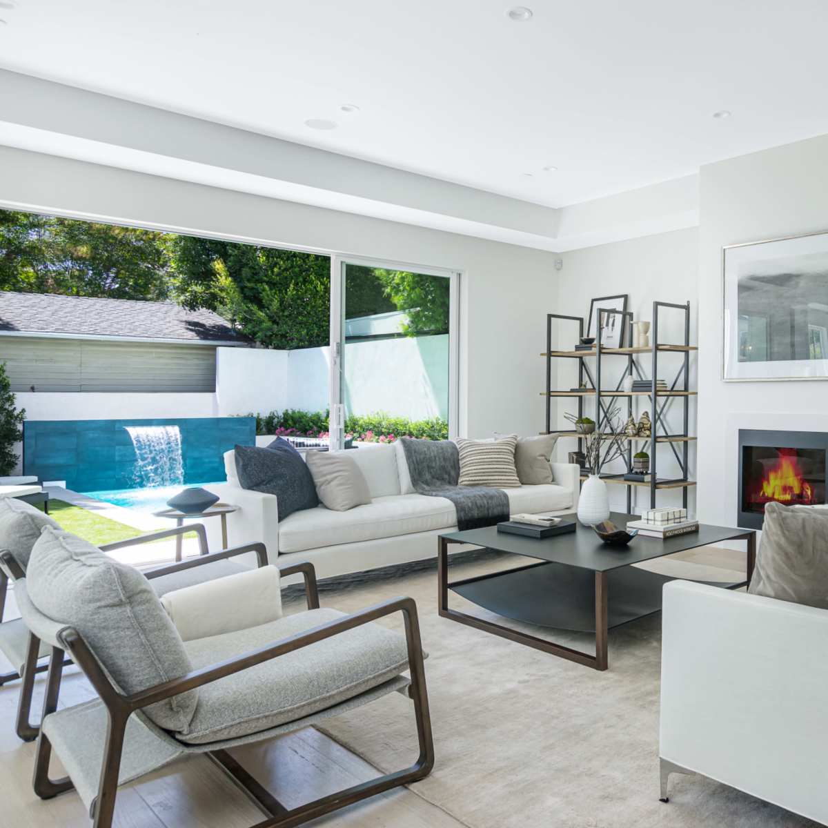 A modern living room with light-colored furnishings, a central coffee table, and large windows overlooking a pool area.