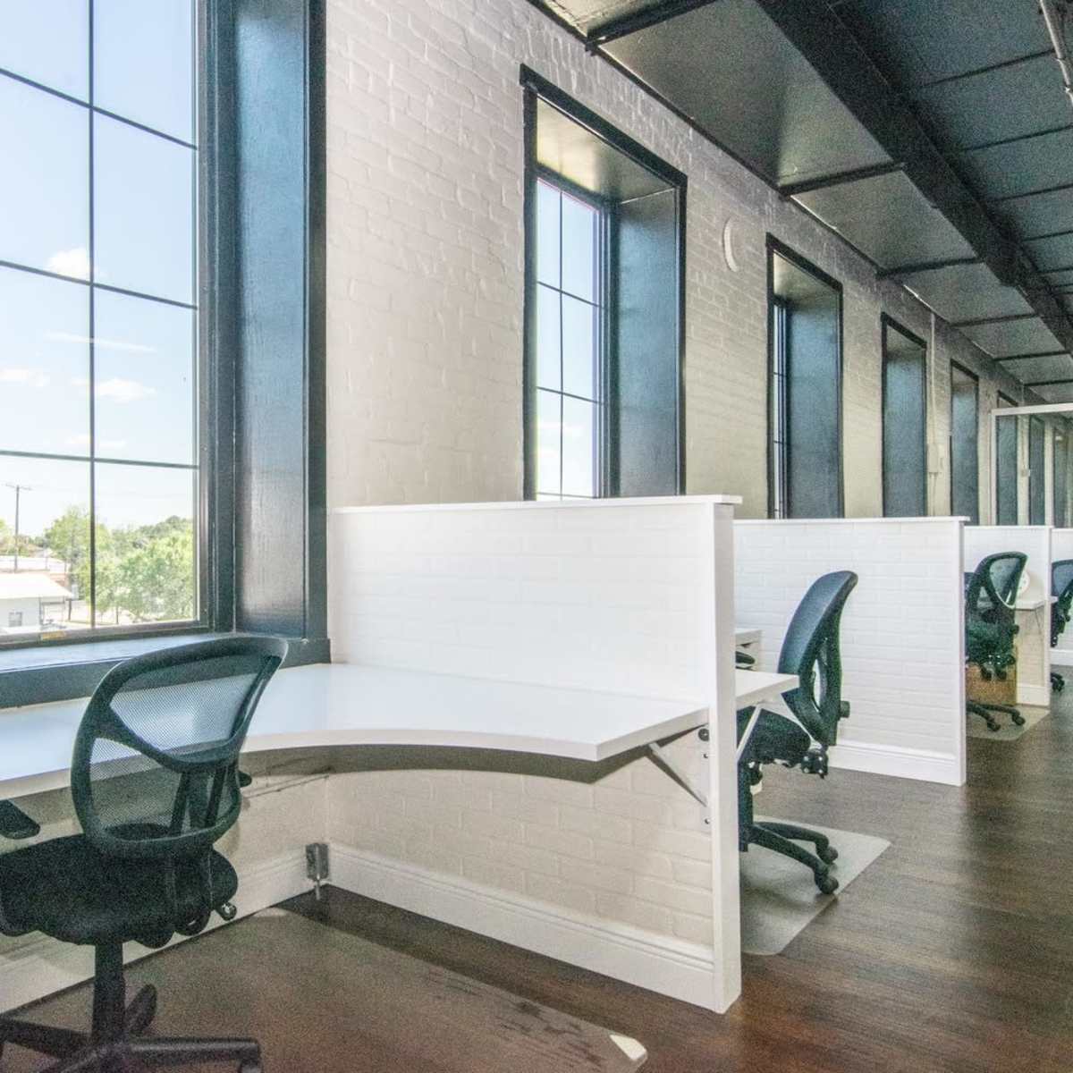 A modern office space with large windows, white walls, and a series of white desks arranged against the walls.