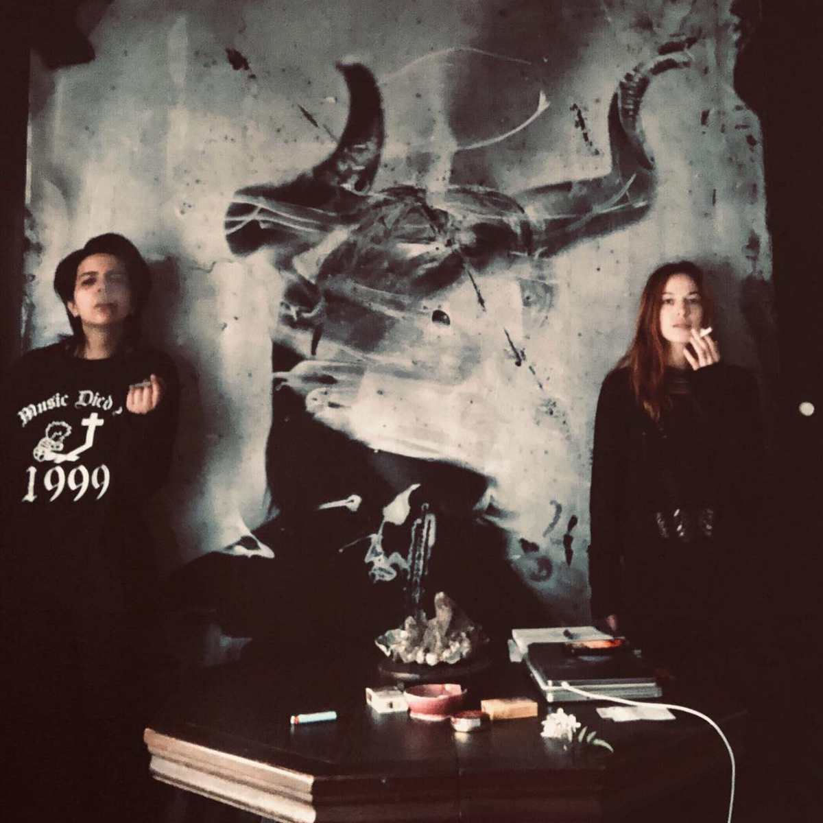 Two individuals stand beside a large, dark wall mural depicting a bull skull, with a table in front holding various items.