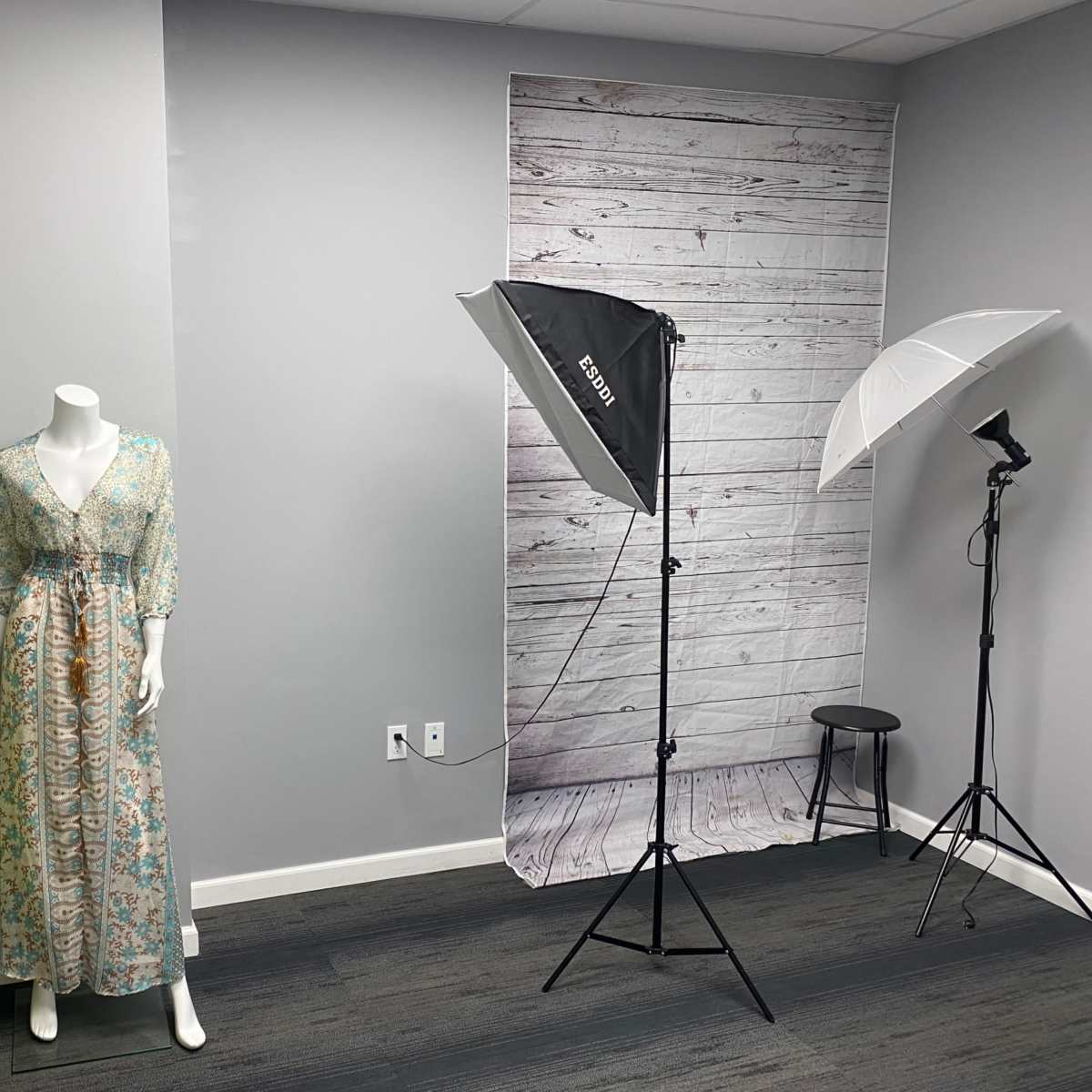 A mannequin wearing a floral dress stands next to photography lighting equipment in a room with gray walls and a wood-patterned backdrop.