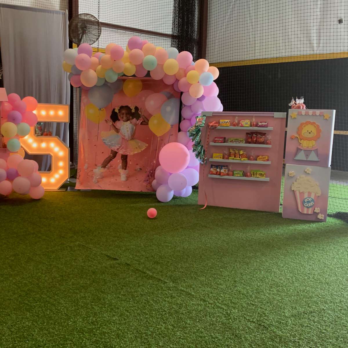 A brightly decorated indoor party area featuring a pink backdrop with balloons, a cart with snacks, and colorful shelves displaying treats.