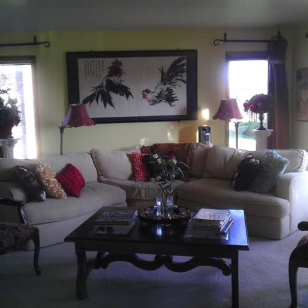 A cozy living room with a light-colored sectional sofa, a dark wooden coffee table, decorative pillows, and large windows allowing natural light to enter.