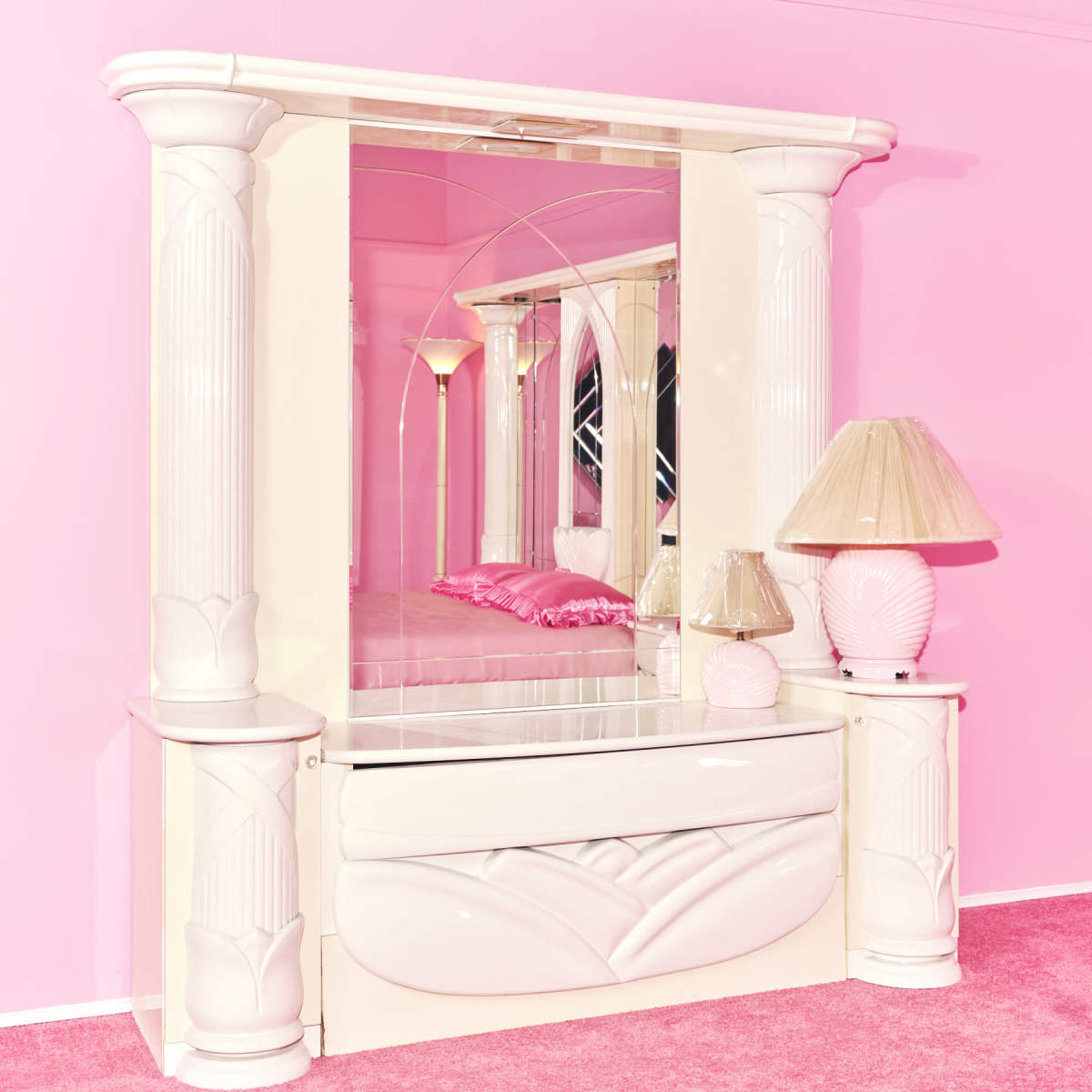 A white dresser with a large mirror and decorative columns, set against a pink background and accompanied by a pink lamp.