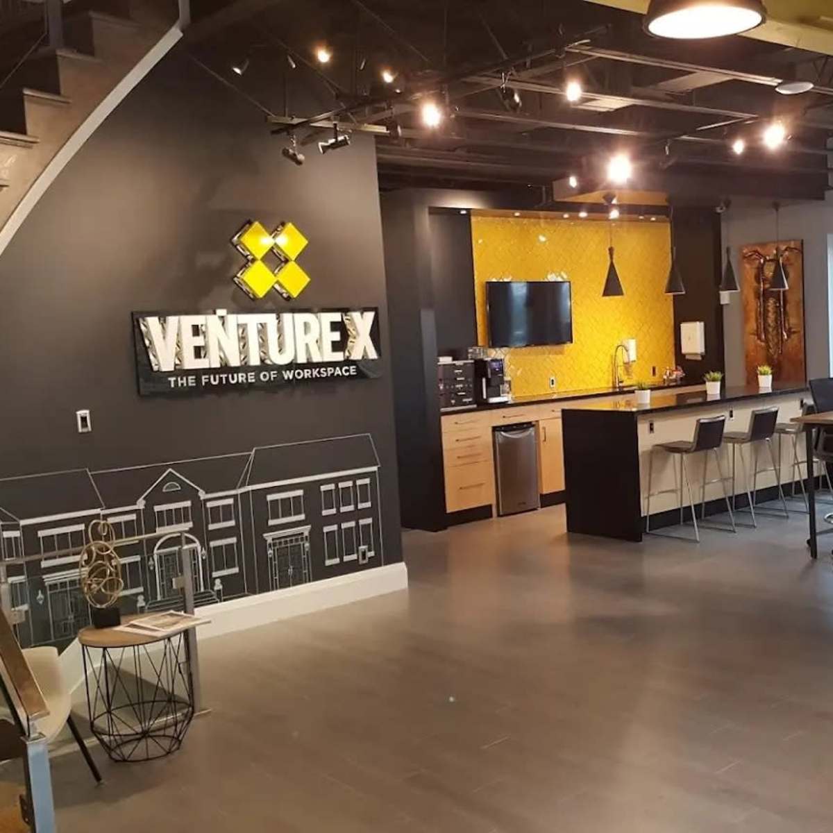 A modern office space featuring a reception area with a prominent "Venture X" sign, a kitchen counter, and various workstations.